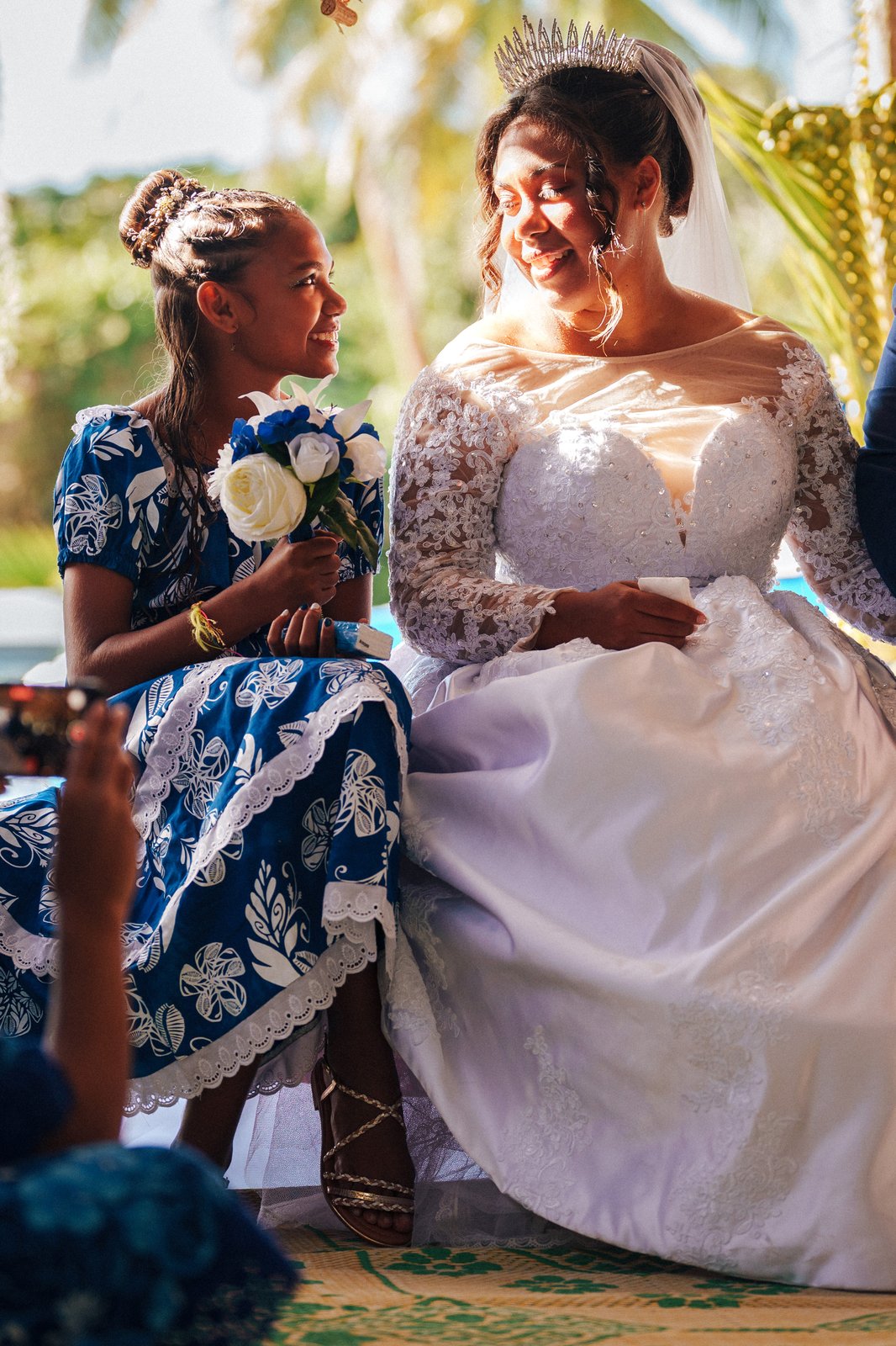 Sous les palmiers, la mariée partage un sourire complice avec une jeune demoiselle d’honneur — Timi M. Bourdet