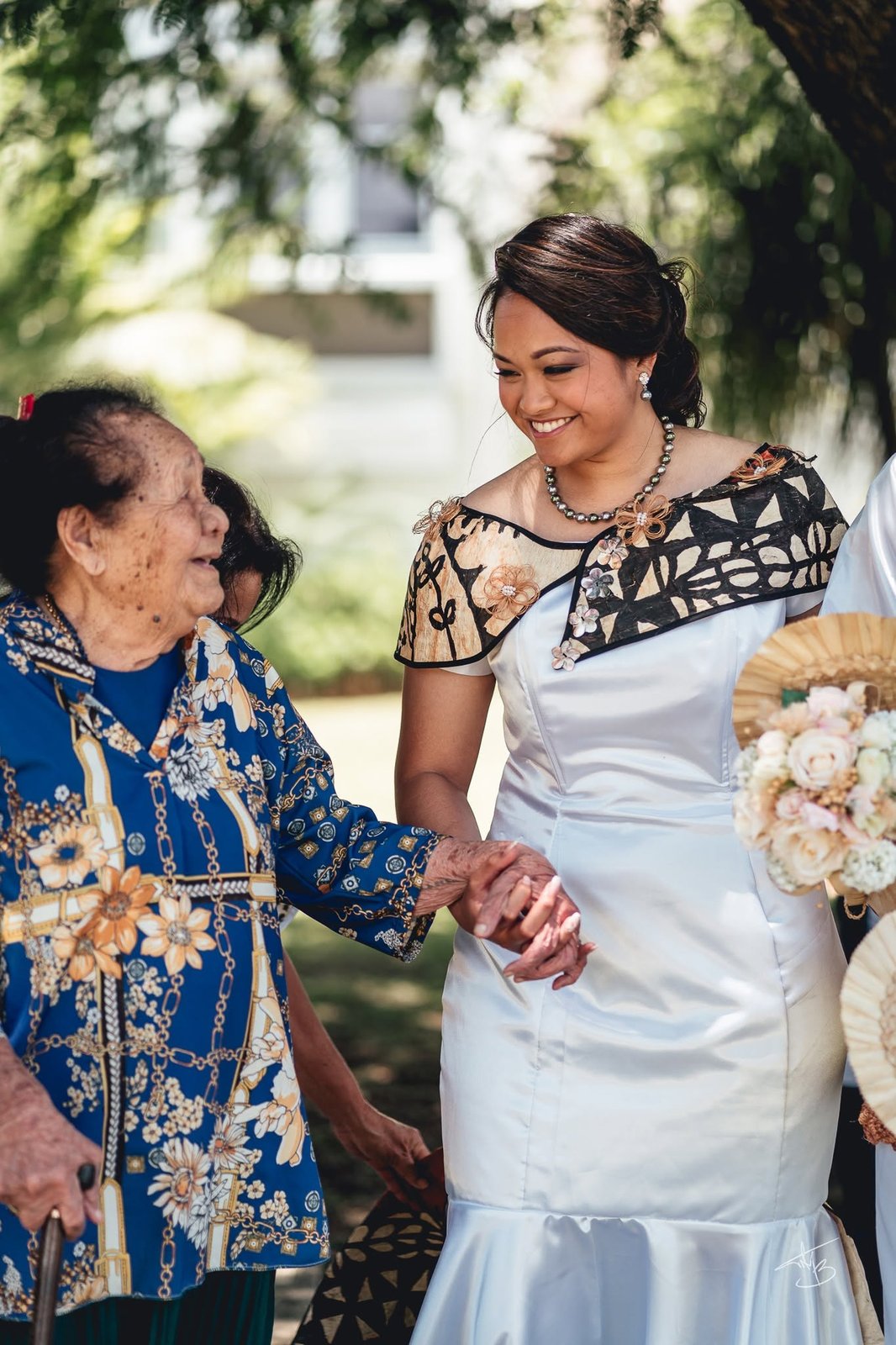La mariée en robe ornée de tapa tient la main d’une aînée souriante, à l’ombre du jardin — Timi M. Bourdet