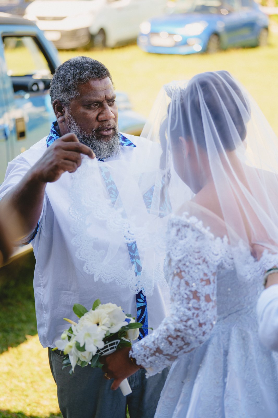 Geste tendre avant la cérémonie : un proche en chemise océanienne ajuste le voile de la mariée — Timi M. Bourdet