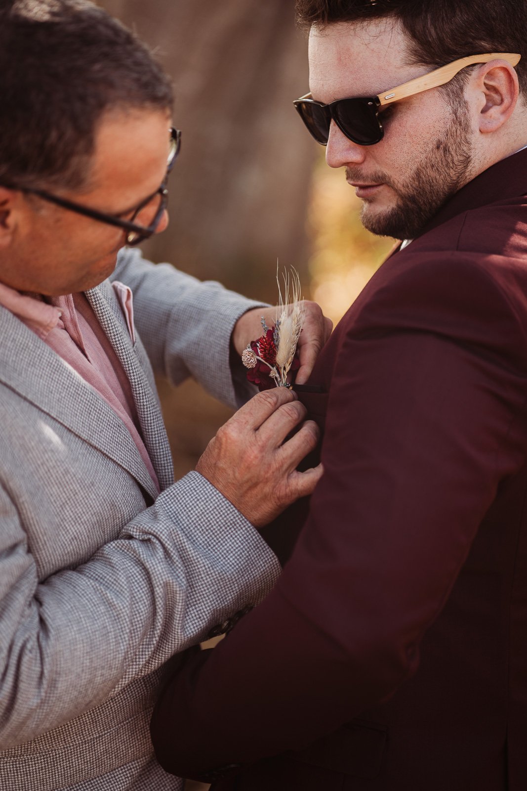 Un proche épingle la boutonnière de fleurs séchées au revers du marié — Timi M. Bourdet